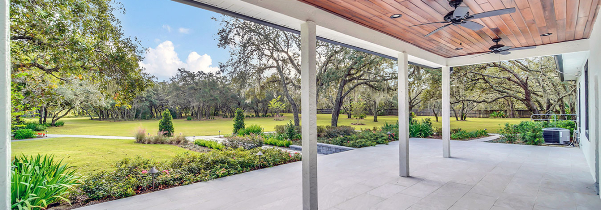 Covered patio with wood ceiling, ceiling fans, and stone tile flooring overlooking a landscaped lawn, garden beds, and mature trees in Orlando, Florida