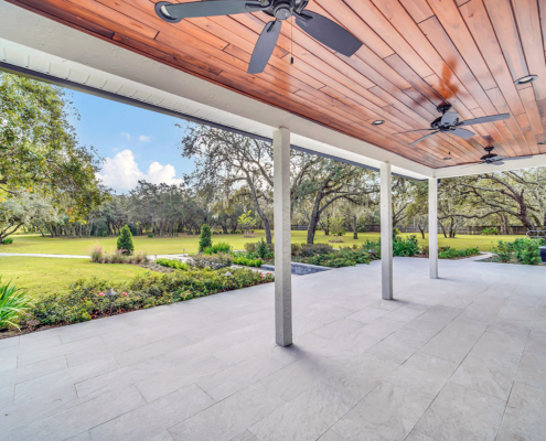Covered patio with wood ceiling, ceiling fans, and stone tile flooring overlooking a landscaped lawn, garden beds, and mature trees in Orlando, Florida
