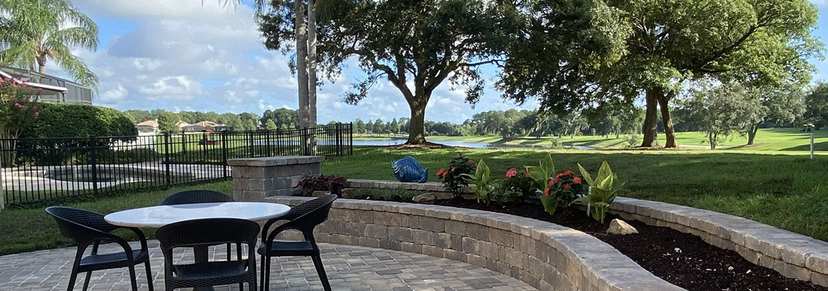 Paver patio with outdoor dining furniture, curved stone planter walls, mature trees, and a lakefront view in the Orlando Florida area.