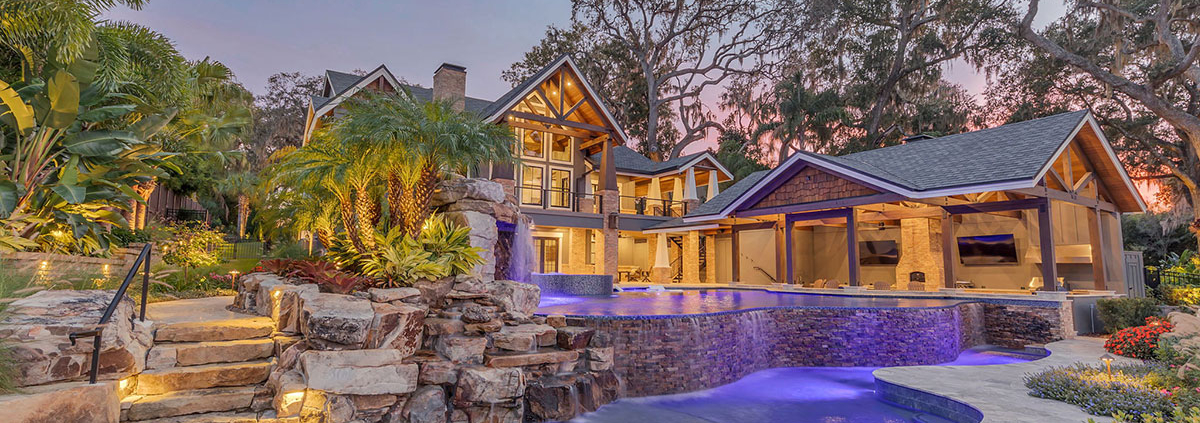 Luxury Orlando estate backyard featuring a multi-level swimming pool, natural stone waterfalls, tropical landscaping, illuminated stone steps, and a covered outdoor living pavilion.