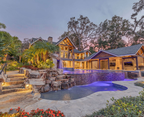 Luxury Orlando estate backyard featuring a multi-level swimming pool, natural stone waterfalls, tropical landscaping, illuminated stone steps, and a covered outdoor living pavilion.