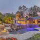 Luxury Orlando estate backyard featuring a multi-level swimming pool, natural stone waterfalls, tropical landscaping, illuminated stone steps, and a covered outdoor living pavilion.